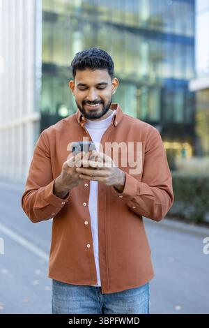 A smiling man uses his smartphone outdoors, likely checking social media or messages, near a modern building. Stock Photo