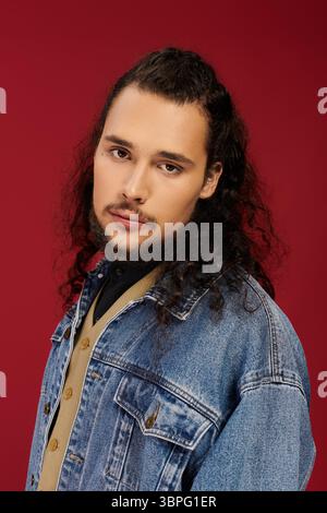 A charismatic young bearded man poses in a denim jacket against a striking red background. Stock Photo