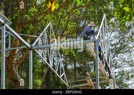 A worker welds a steel frame on a high scaffold surrounded by trees, merging modern construction with nature Stock Photo