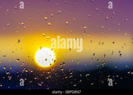 A dreamy sunset glows through raindrops on glass, blending golden light and purple hues in a serene and moody evening scene Stock Photo