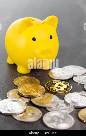 A yellow piggy bank surrounded by scattered gold and silver Bitcoin coins on a dark surface, representing cryptocurrency saving and financial concepts Stock Photo