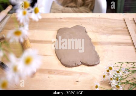 Clay workshop setup with rolled-out clay, rolling pin, tools, and fresh chamomile flowers on a wooden table. Cozy atmosphere and natural materials in Stock Photo