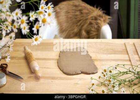 Clay workshop setup with rolled-out clay, rolling pin, tools, and fresh chamomile flowers on a wooden table. Cozy atmosphere and natural materials in Stock Photo