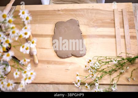 Clay workshop setup with rolled-out clay, rolling pin, tools, and fresh chamomile flowers on a wooden table. Cozy atmosphere and natural materials in Stock Photo