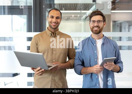 Two diverse colleagues smile, holding a laptop and tablet, in a modern office setting. They look friendly and professional. Stock Photo