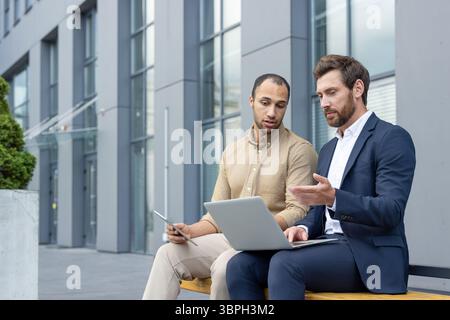 Two business professionals collaborating outdoors near a modern building, using a laptop and a tablet. Stock Photo