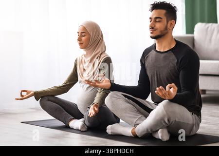 Expecting Muslim Spouses Practicing Yoga Together At Home, Sitting In Lotus Pose Stock Photo