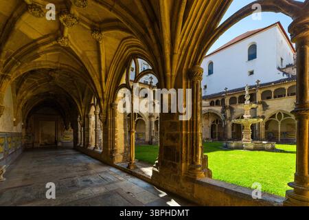 view of a sunlit Gothic cloister, featuring long vaulted arcades, a central green courtyard with a fountain, and a bright white architectural building Stock Photo