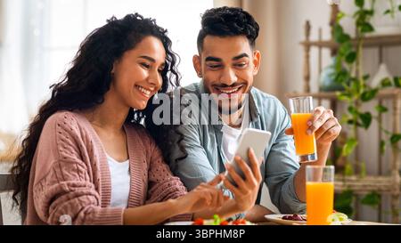 Middle eastern couple watching photos on smartphone while having breakfast in kitchen Stock Photo