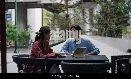 Two businesspeople are discussing work at an outdoor cafe, using a tablet and enjoying coffee Stock Photo