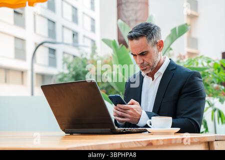 A dedicated businessman in a sharp suit is intently using his laptop and smartphone while working remotely at a terrace cafe, showcasing a blend of Stock Photo