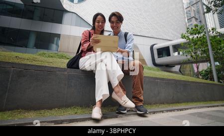 Professionals collaborating outdoors, checking digital tablet, sharing smile near contemporary cityscape Stock Photo