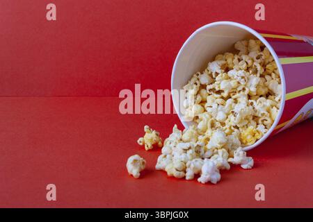 Popcorn spills out of a striped paper bucket on a red background, copy space Stock Photo