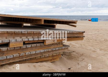 Stacked wooden boardwalks lie on a sandy beach, with the sea in the background and a blue beach hut in the distance. Stock Photo