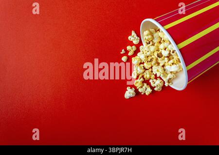 Popcorn spills out of a striped paper cup on a red background, copy space Stock Photo