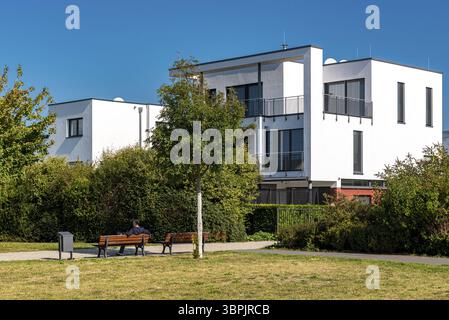 A modern house in an upscale living style with windows, balcony and terrace and a park with park benches in the foreground under a cloudless sky, Fran Stock Photo