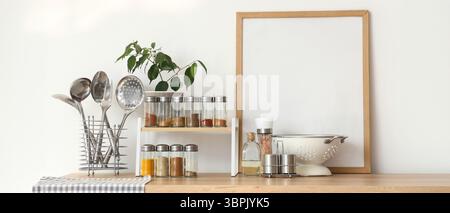 Shelf with different spices, plant, frame and utensils on counter near light wall in kitchen Stock Photo