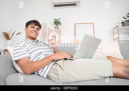 Handsome young man with air conditioner remote control using laptop on sofa at home Stock Photo