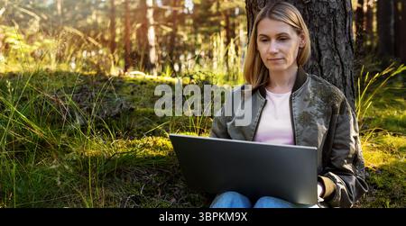 woman using laptop in the park. digital nomad sitting against the tree and work remotely and enjoy the nature. freelance remote working concept. banne Stock Photo