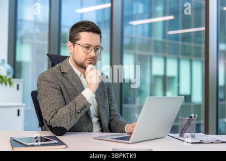 Serious young male businessman sitting at his desk in the office, looking thoughtfully at the laptop screen. Stock Photo