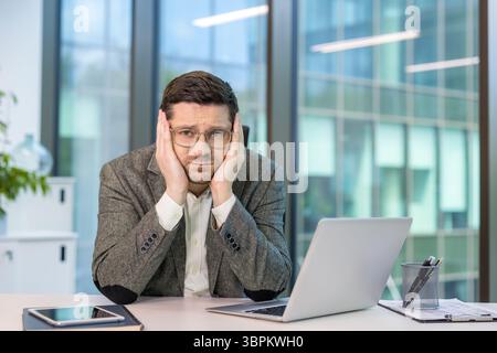 Close-up portrait of a young male office worker sitting at his desk, head resting on his hands, looking bored and tired at the camera. Stock Photo