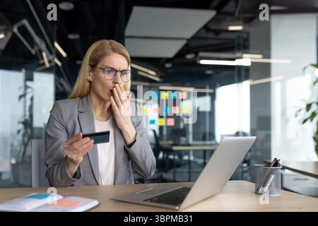 A businesswoman reacts in shock while looking at a credit card and a laptop. She seems surprised at the result of her transaction. Stock Photo