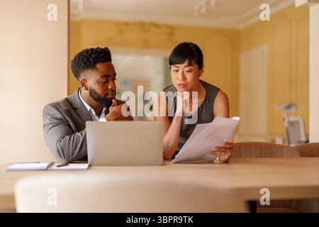 Focused professionals discussing project details and reviewing documents with laptop on desk in office Stock Photo