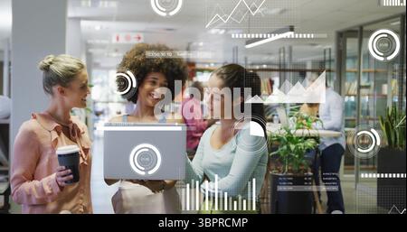 Standing three businesswomen collaborating over silver laptop in open plan office, with coffee cup Stock Photo