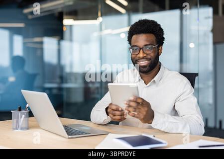 A smiling African American man in glasses uses a tablet at his office desk, a laptop sits nearby. Stock Photo