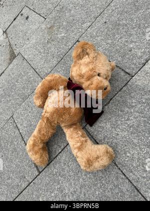 The image displays a light brown plush teddy bear with a dark red velvet bow lying on its side on a grey stone pavement made of interlocking tiles. Th Stock Photo