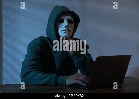Anonymity. Masked man with laptop showing hush gesture at table in dark Stock Photo