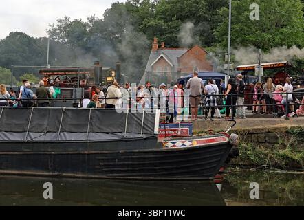 Former working boats are moored along the banks of the Bridgewater canal in front of the smoking traction engines in town for the Lymm transport day Stock Photo