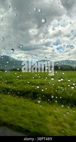 View from inside a car showing raindrops on the window, with blurred green meadows and stormy sky in the background. Summer countryside road trip mood Stock Photo