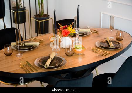 Elegantly set wooden oval dining table in a modern dining room. The table is prepared for a festive dinner party with dark plates, gold cutlery, and s Stock Photo