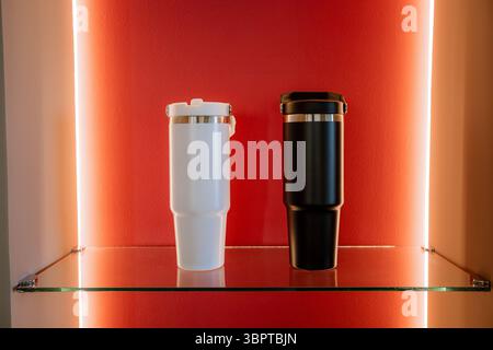 Two minimalist travel tumblers, one white and one black, displayed on glass shelf against illuminated red wall. Mockup Stock Photo