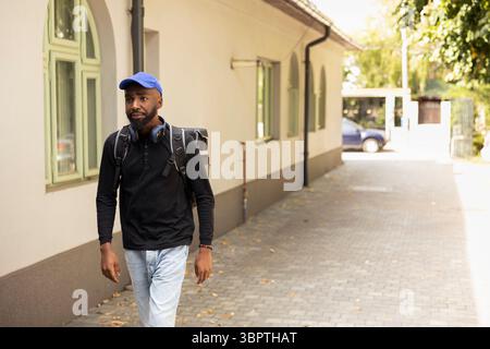 African american courier with thermal backpack approaches a doorway to deliver a food takeaway order. Using a smartphone app, he confirms the drop off location and rings the intercom. Stock Photo