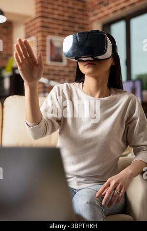 Self employed woman wearing vr goggles, seated on couch, focusing on virtually preparing business presentations. Asian female freelancer exploring augmented reality tools for career and productivity. Stock Photo