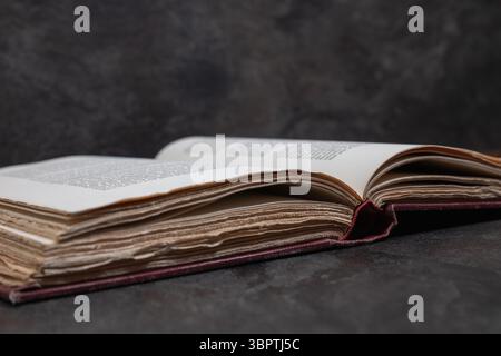 Close side view of an open antique book, focus on deckled page edges, resting on dark textured surface with moody background and angled perspective Stock Photo