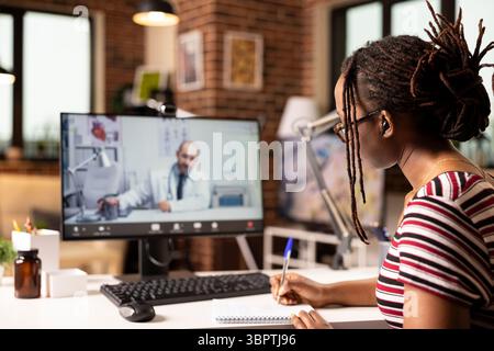 Female patient sits at home in front of computer, having online medical consultation with remote doctor via video call. Black woman taking notes in notebook while speaking to physician on desktop PC. Stock Photo