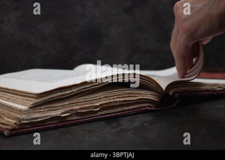 Open antique book lying on dark surface with dark background, a hand flipping pages showing the back of the hand, shallow focus hides text. Stock Photo