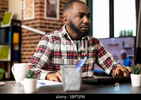 Focused black man working from home, reviewing company records on desktop pc. African american entrepreneur with hands on keyboard and mouse, updating business fiscal reports before deadline. Stock Photo