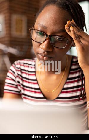 Female entrepreneur experiences headache while seated at home office desk, trying to manage multiple projects. Close up of fatigued woman massaging her temples, struggling to focus on laptop. Stock Photo