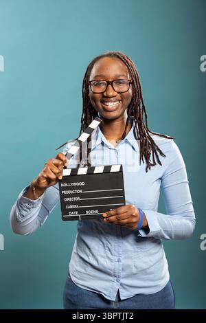 Portrait of african american female movie director happily holding clapperboard, ready for shooting. Smiling female filmmaker poses with film slate in hand, looking at camera with enthusiasm. Stock Photo