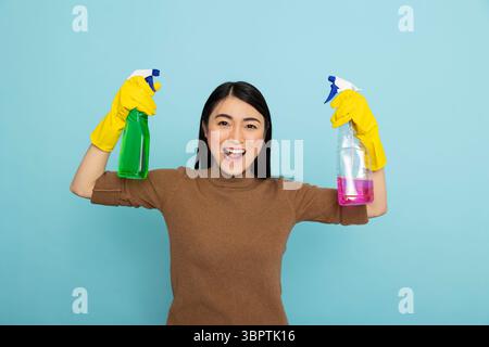 Energetic asian housekeeper in rubber gloves holding colorful cleaning sprays and flexing arms, promoting cleanliness with big smile. Enthusiastic female cleaner with pink and green liquid solutions. Stock Photo