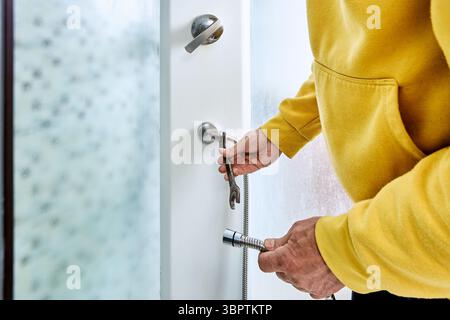 Using open end wrench to fasten metal nut, plumber secures one end of shower hose to plumbing connector on vertical wall surface. Stock Photo