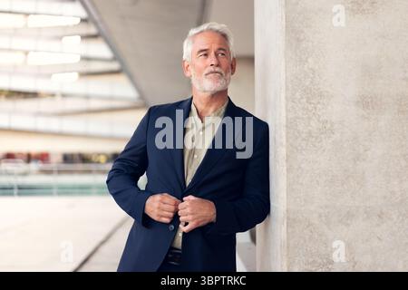 Senior businessman in formal outfit standing outdoors near concrete wall, buttoning blazer. Calm and thoughtful expression in modern urban setting Stock Photo