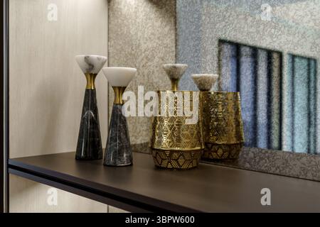 Elegant home decor with marble candle holders and a gold patterned vase on a dark shelf. Luxurious interior design detail against a textured mirror wa Stock Photo