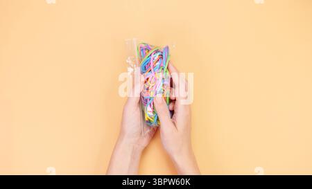 Top view of hands holding a clear plastic bag filled with colorful rubber bands on a beige background. Perfect for crafting, DIY projects, and organiz Stock Photo
