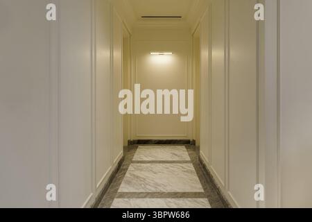 Long, elegant hallway with classic white wall molding and a luxurious polished marble floor. A single modern light fixture illuminates the perspective Stock Photo