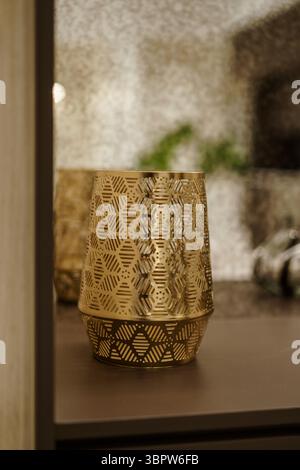 A close-up of an elegant gold metal candle holder with an intricate geometric perforated pattern, sitting on a dark shelf with a soft, blurred backgro Stock Photo